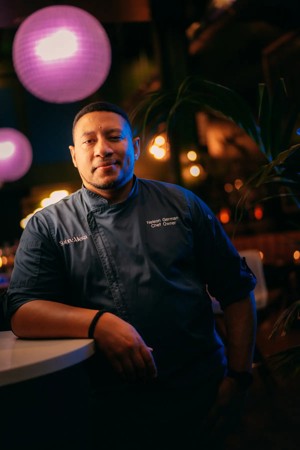Chef Nelson German standing beneath warm pendant lights inside Sobre Mesa